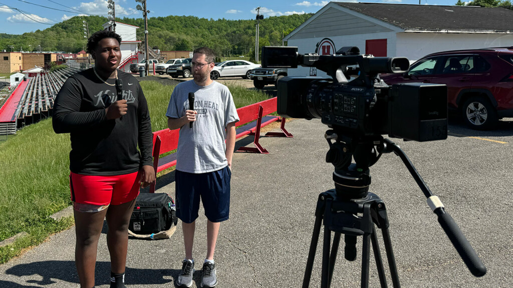 Da'Ron Parks and Marc Givler speaking with microphones in their hands overlooking the football stadium at Hurricane high school in West Virginia. 